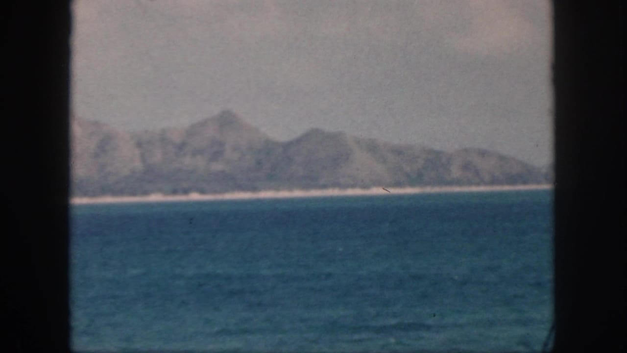 1959 Oahu home-movie frame showing calm ocean water, a pale shoreline, and distant rugged mountains under a hazy sky.
