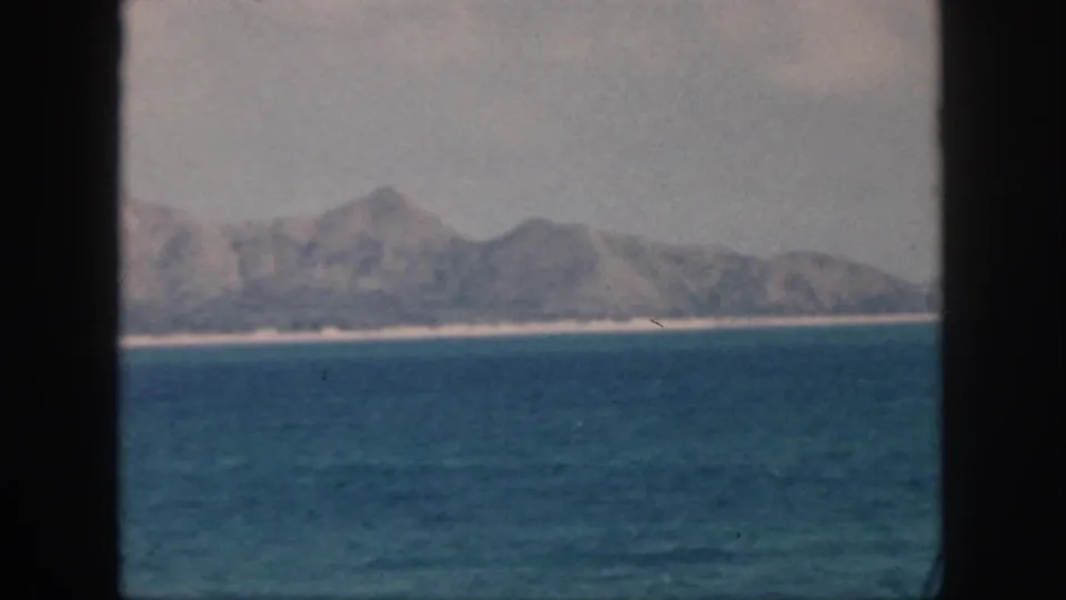 1959 Oahu home-movie frame showing calm ocean water, a pale shoreline, and distant rugged mountains under a hazy sky.