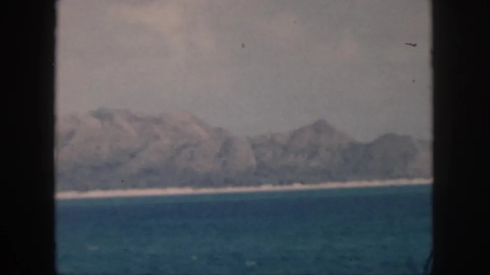 1959 Oahu home-movie frame with a tilted horizon, calm water, pale shore, and rugged mountains softened by haze and film grain.