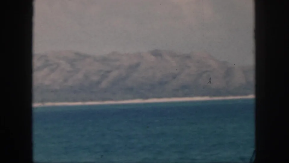 1959 Oahu footage frame showing a broad blue ocean foreground, thin coastal strip, and hazy mountains beyond the water.