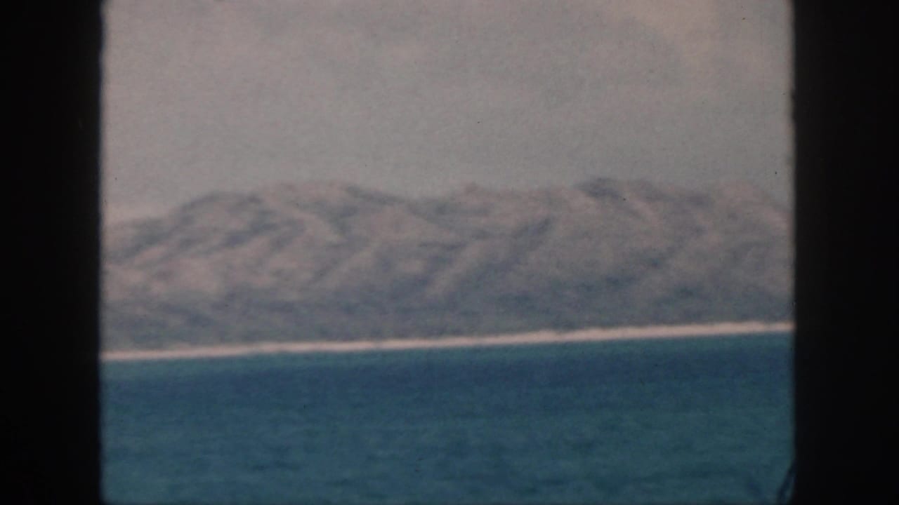 1959 Oahu-area coastline viewed from water through dark framing, with mountains, pale shoreline, and hazy sky.