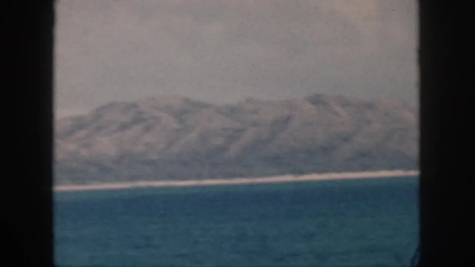 1959 Oahu-area coastline viewed from water through dark framing, with mountains, pale shoreline, and hazy sky.