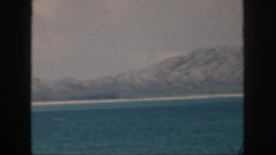 Faded 8mm view of a 1959 Oahu-area coast, with turquoise water, pale shore, and hazy mountains beyond.