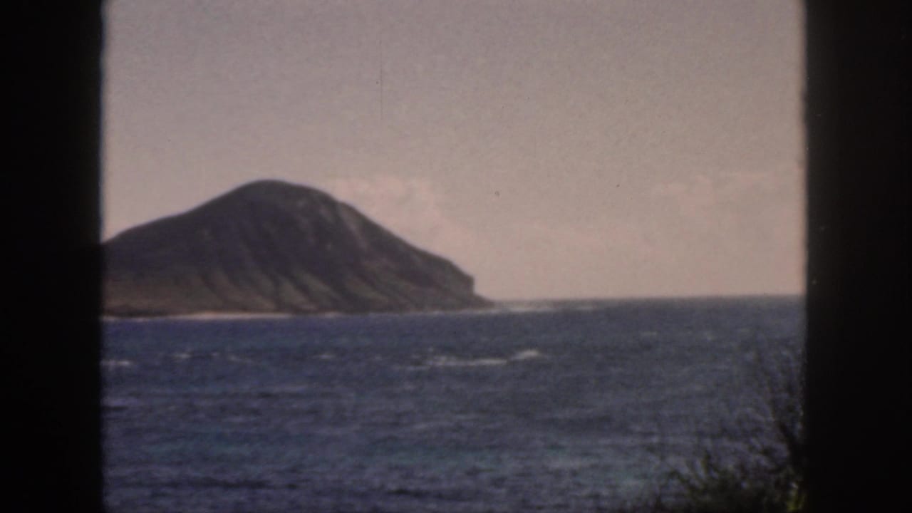 1959 Oahu 8mm frame showing a ridged offshore island across blue water, seen through dark vertical framing edges.