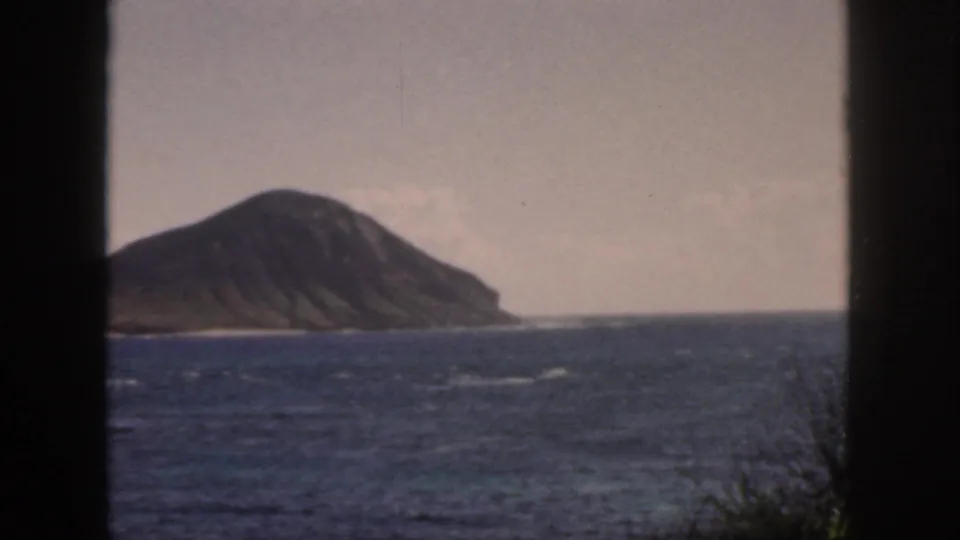 1959 Oahu 8mm frame showing a ridged offshore island across blue water, seen through dark vertical framing edges.