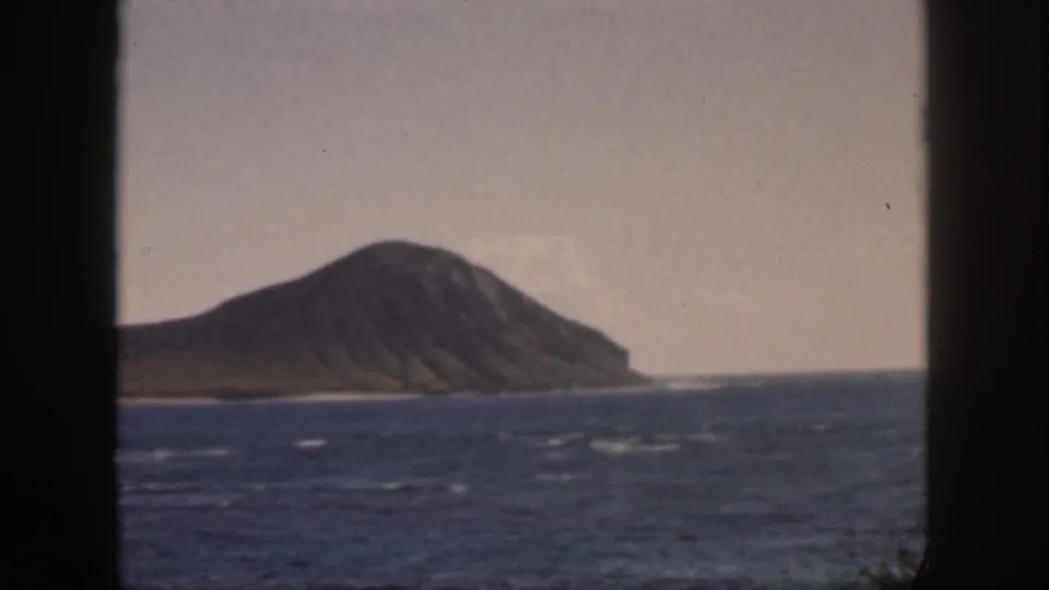1959 Oahu home-movie frame with an offshore island, pale sky, and choppy blue water framed by dark window or doorway edges.