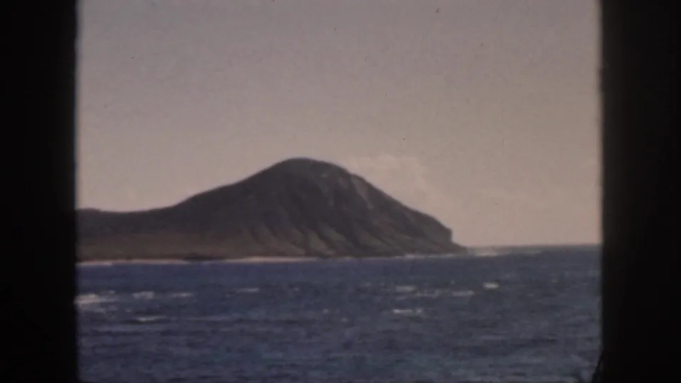 1959 Oahu 8mm frame showing the ridged island and shoreline band across blue ocean, with dark framing at left and right.