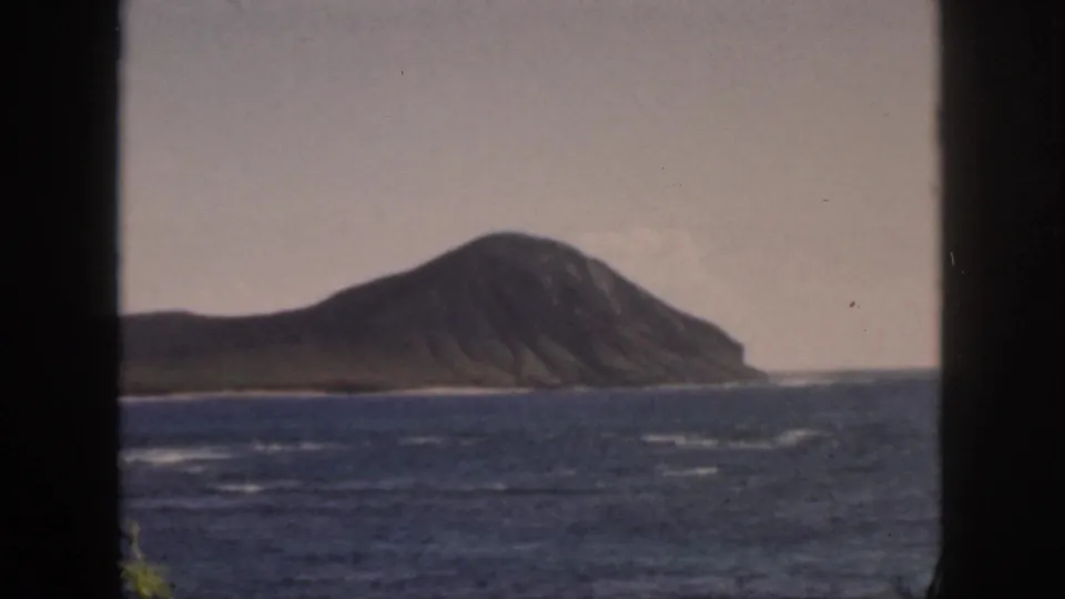 1959 Oahu home-movie frame of a volcanic-looking island across the ocean, framed by dark vertical edges and soft film grain.