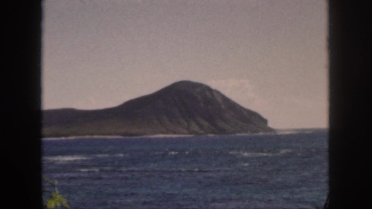 1959 Oahu home-movie view of a large volcanic island across choppy blue water, seen through dark window or doorway edges.