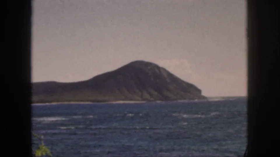 1959 Oahu home-movie view of a large volcanic island across choppy blue water, seen through dark window or doorway edges.