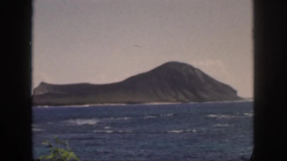 1959 Oahu 8mm frame with a sloped volcanic island across blue water and faint foreground greenery near the lower edge.