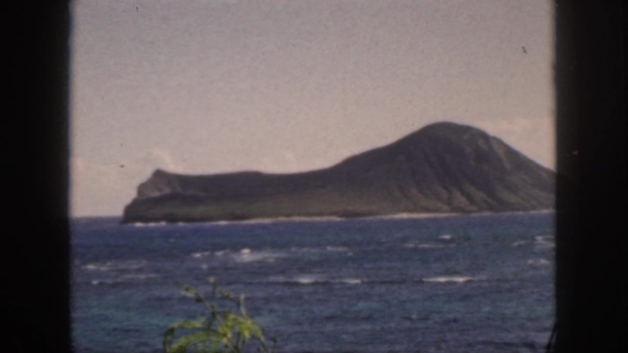 1959 Oahu home-movie view of a distant rugged island across blue, wind-ruffled ocean, seen through dark foreground framing.