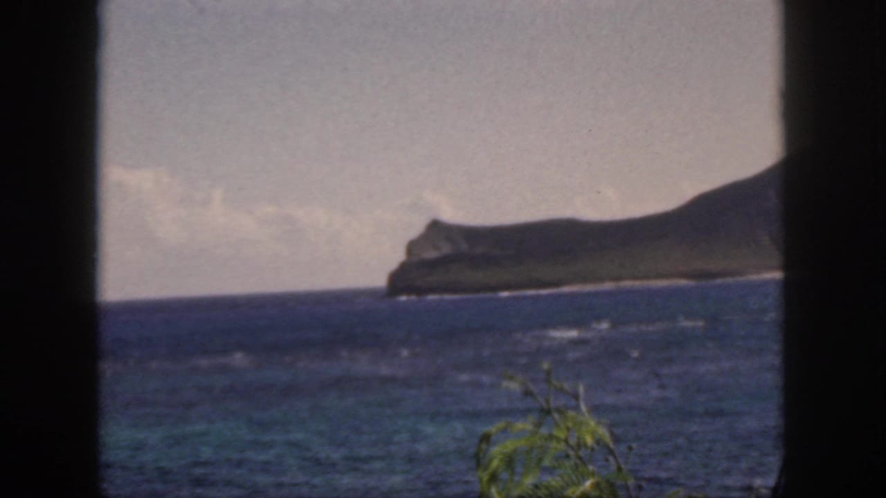 1959 Oahu 8mm frame showing blue ocean, whitecaps, a distant green coastal headland, and foliage seen through a dark window-like frame.