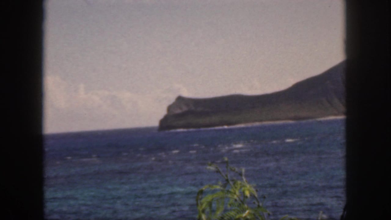 1959 Oahu home-movie frame showing a distant island beyond blue water, with dark window-like framing and foliage at the lower edge.