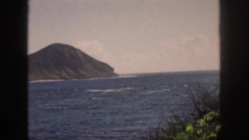 1959 Oahu home-movie view of a rounded offshore headland across the sea, with soft focus, foliage, and dark framing at the sides.