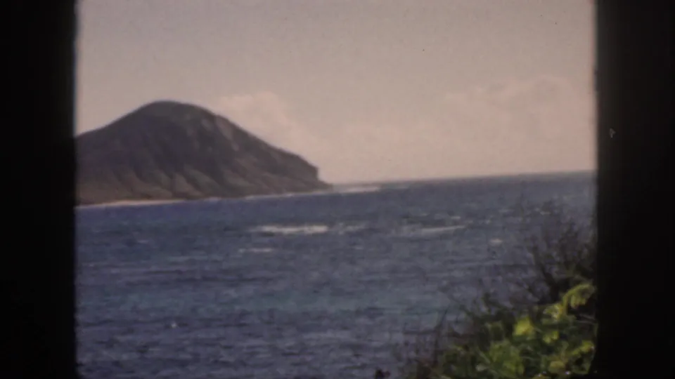 1959 Oahu frame showing a sloped island shoreline beyond blue water, with soft grain, foreground foliage, and dark framing.