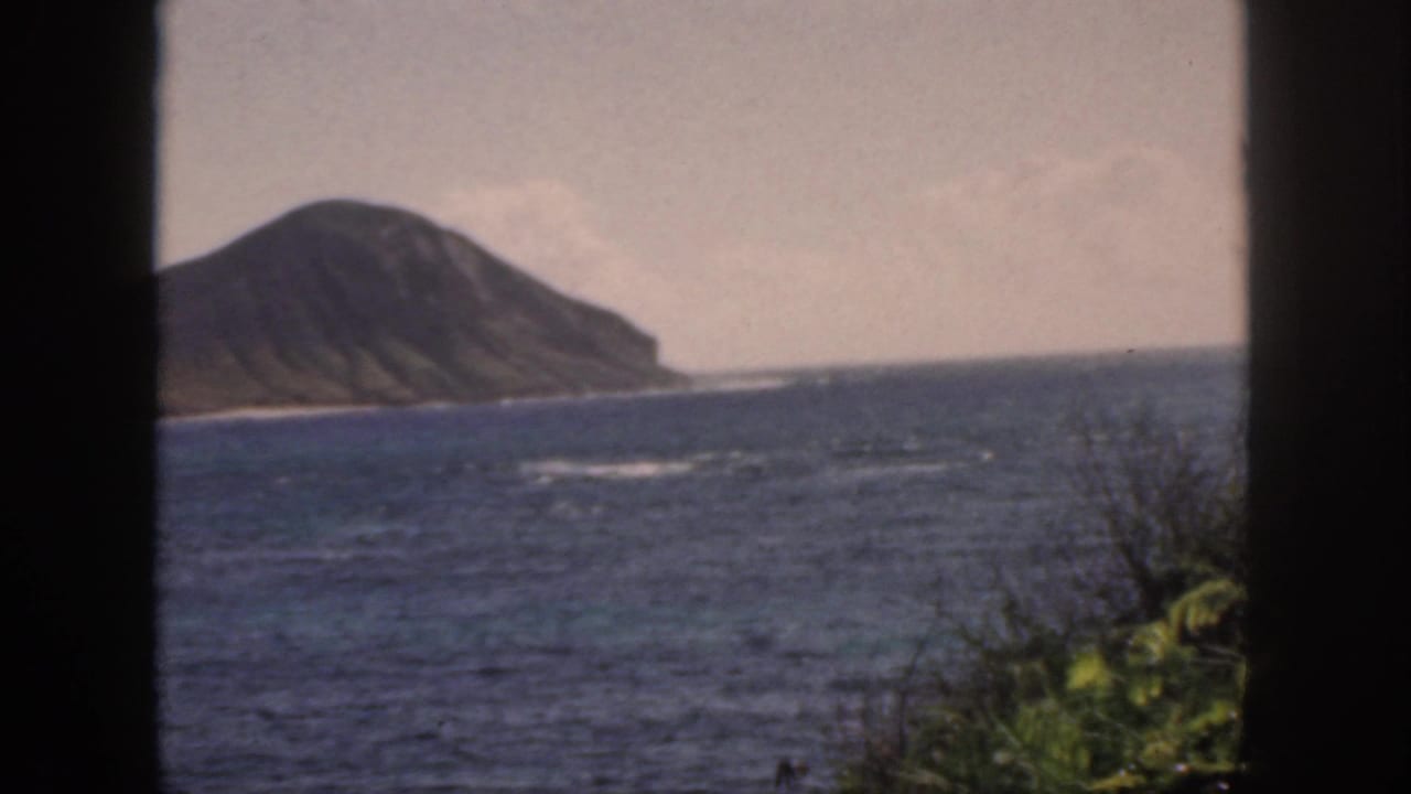 1959 Oahu coastal view with a volcanic landform, deep blue water, hazy sky, and dark vertical window frame in the foreground.