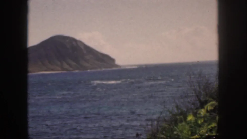1959 Oahu coastal view with a volcanic landform, deep blue water, hazy sky, and dark vertical window frame in the foreground.