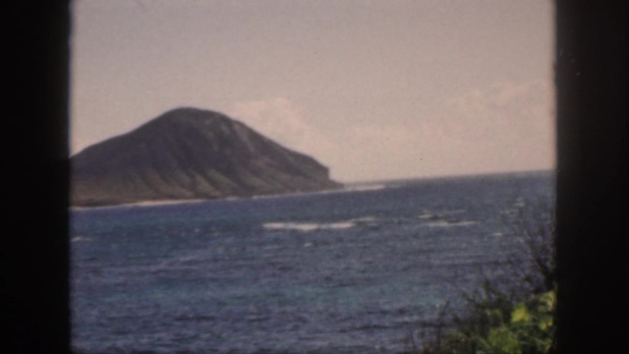 1959 Oahu home-movie frame showing an offshore volcanic landform across blue water, with window bars dividing the view.