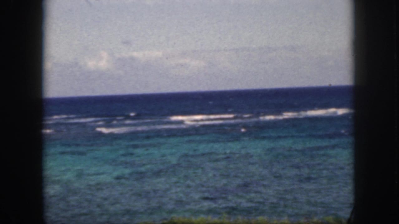 1959 Oahu 8mm frame with offshore surf bands, turquoise water, and a cloudy horizon seen through a dark window-like foreground.
