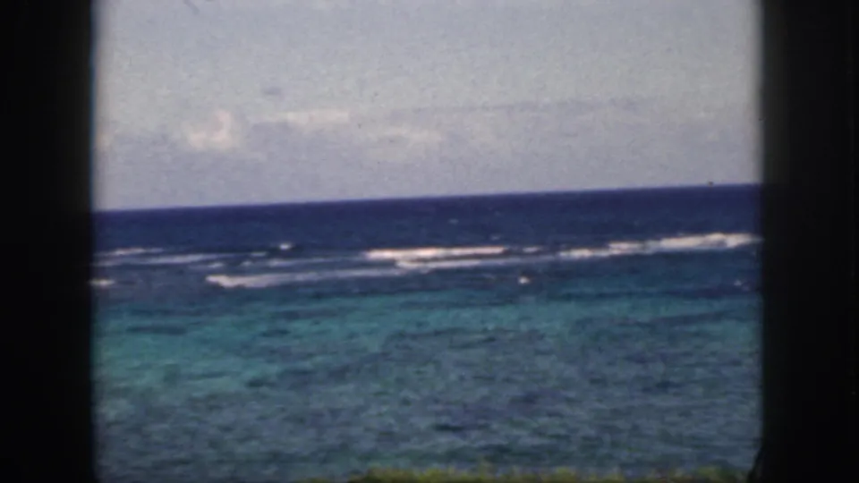 1959 Oahu 8mm frame with offshore surf bands, turquoise water, and a cloudy horizon seen through a dark window-like foreground.