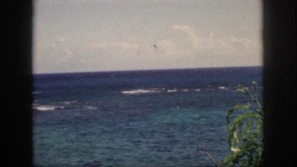 1959 Oahu 8mm frame showing a cloudy tropical ocean view, distant surf breaks, dark foreground bars, and leafy greenery along the lower right edge.