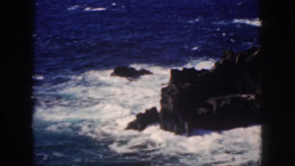 Oahu coastline detail with white Pacific surf breaking against dark volcanic-looking rocks under deep blue water tones.