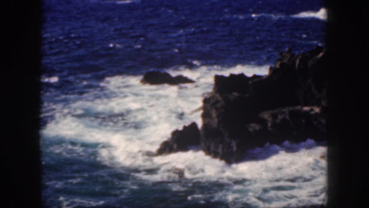 1959 Oahu 8mm frame showing white Pacific surf breaking around dark shoreline rocks.