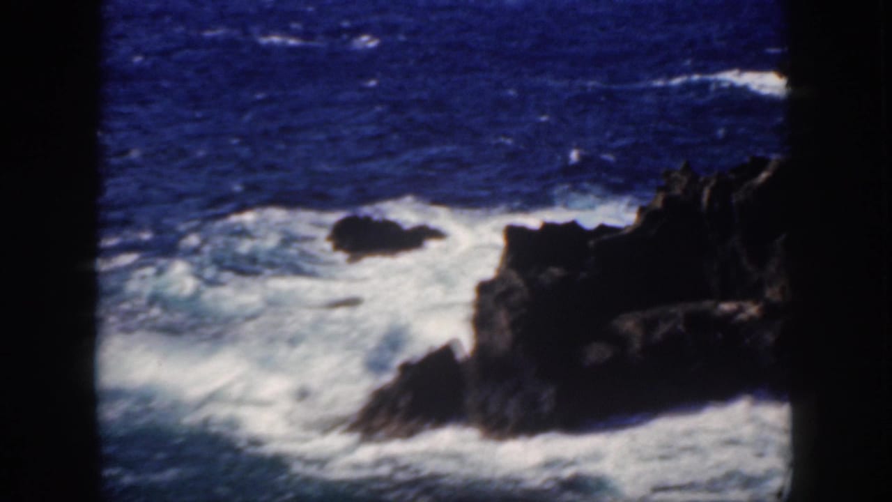 1959 Oahu home-movie frame with surf foaming around a dark rocky shoreline outcrop.