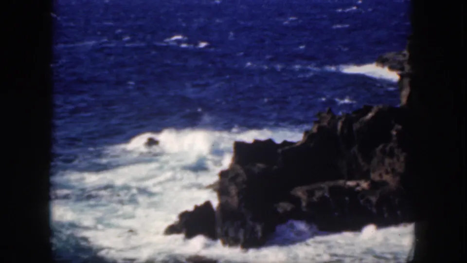Vintage 8mm Oahu frame showing surf rolling into dark rocks on a rugged coast.