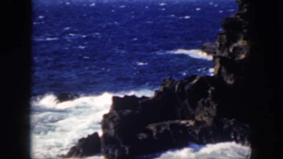 1959 Oahu 8mm frame with white surf wrapping around a dark rocky coastal cliff.