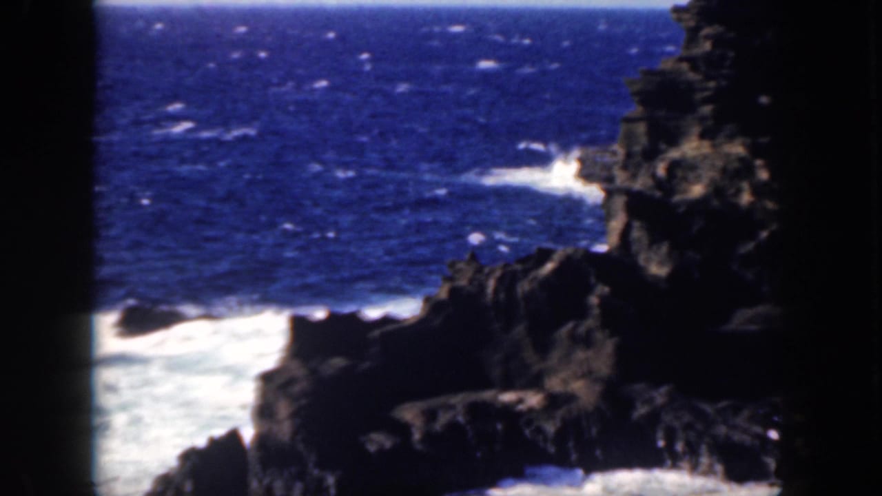1959 Oahu 8mm frame showing jagged volcanic-looking shoreline, blue open water, white surf, and dark frame edges.