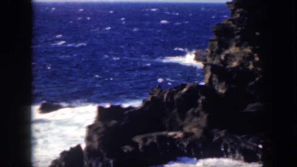 1959 Oahu 8mm frame showing jagged volcanic-looking shoreline, blue open water, white surf, and dark frame edges.