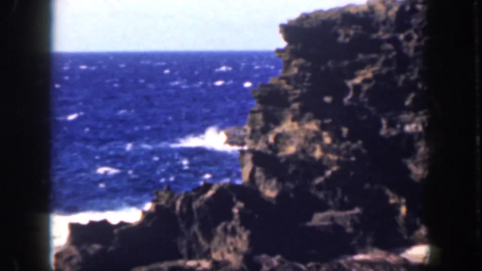 1959 Oahu home-movie frame showing jagged shoreline rocks, bright white surf, blue ocean, and a pale horizon.