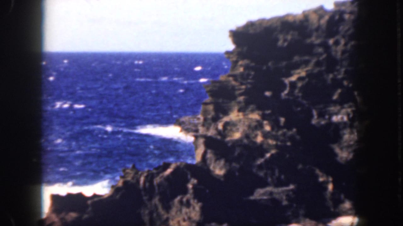 1959 Oahu home-movie frame of deep blue sea and white surf at the base of a dark volcanic-looking cliff.