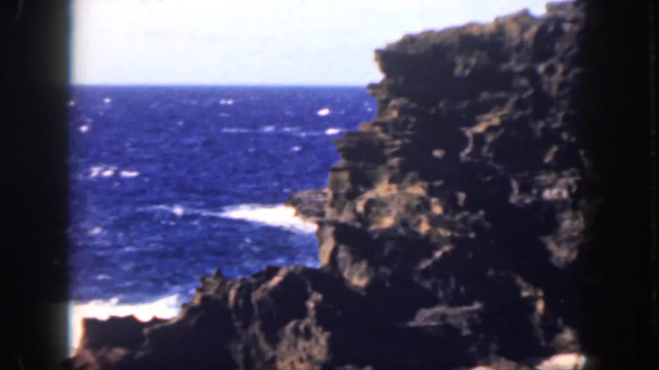 1959 Oahu home-movie frame of deep blue sea and white surf at the base of a dark volcanic-looking cliff.