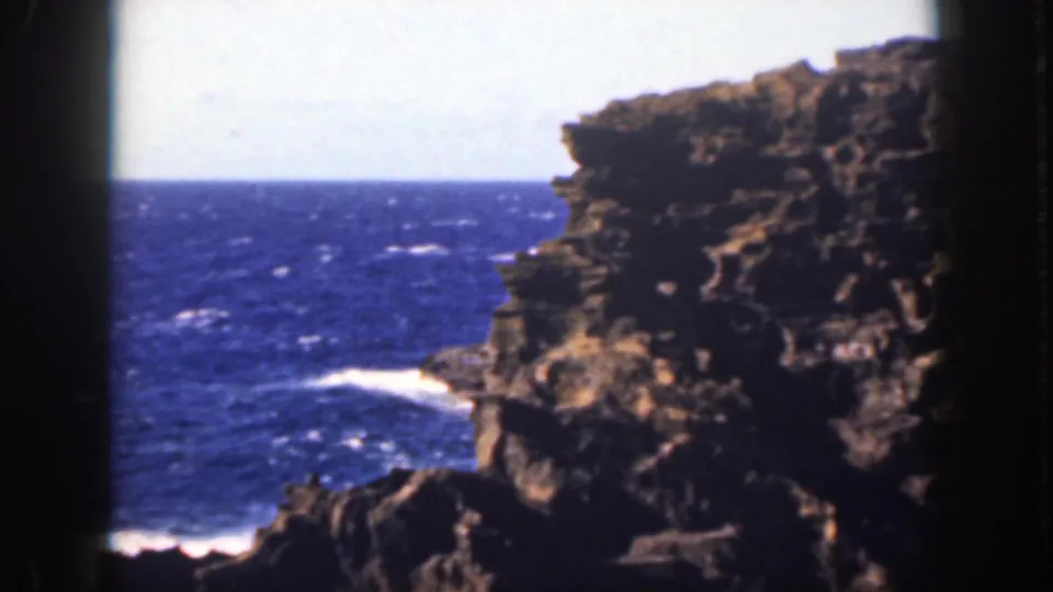 1959 Oahu coastal view with a dark rock face, blue ocean, white breaking waves, and soft 8mm exposure.