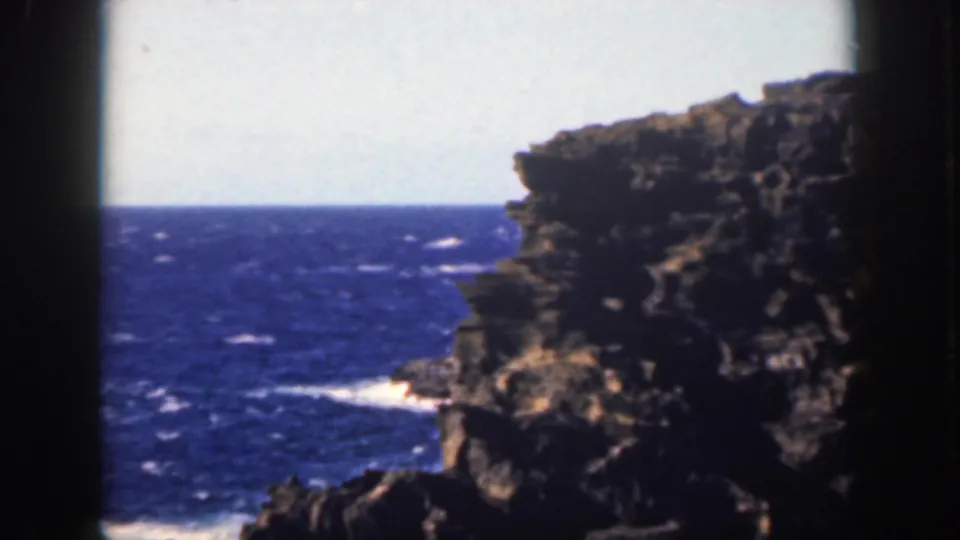 1959 Oahu home-movie view of a rugged ocean cliff with white surf visible below and 8mm gate shadows at the sides.