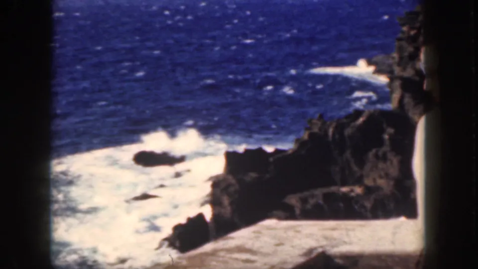 1959 Oahu coastal frame showing foamy surf around dark rocks, with the cliff edge partly framing the right side.