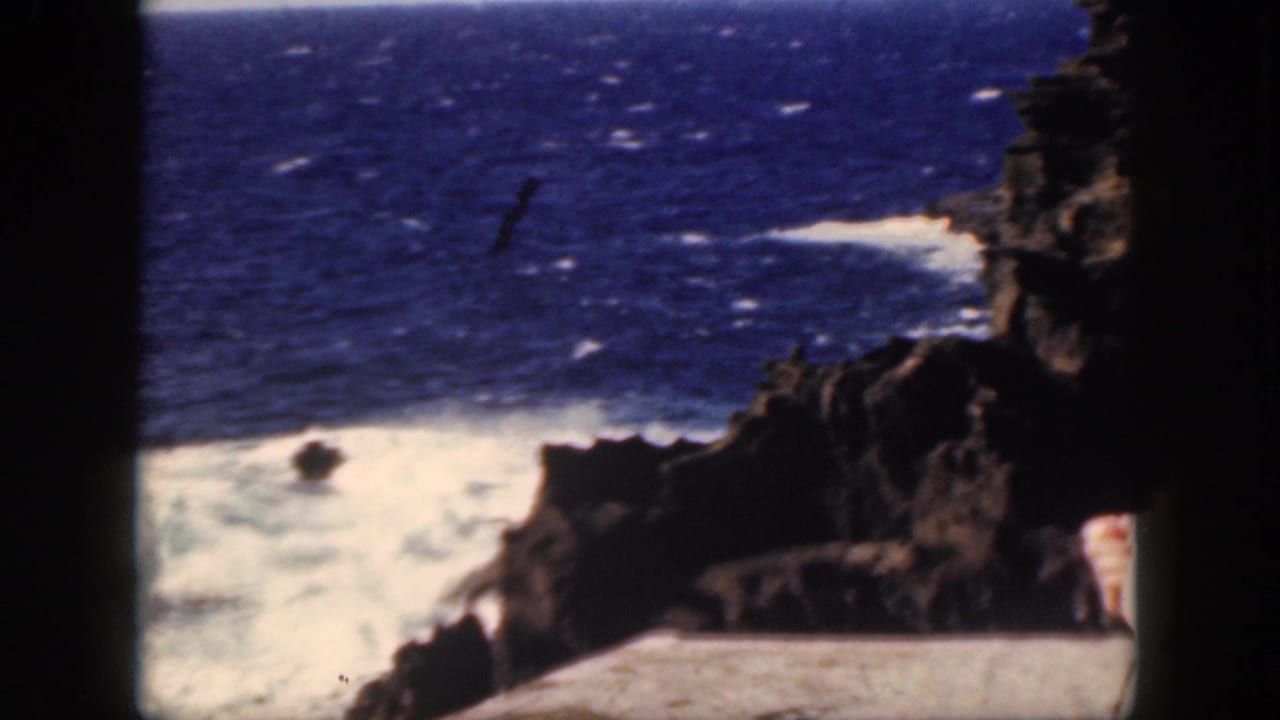 1959 Oahu home-movie frame showing blue ocean, white surf, dark rocks, and a partly visible woman seated near the cliff edge.