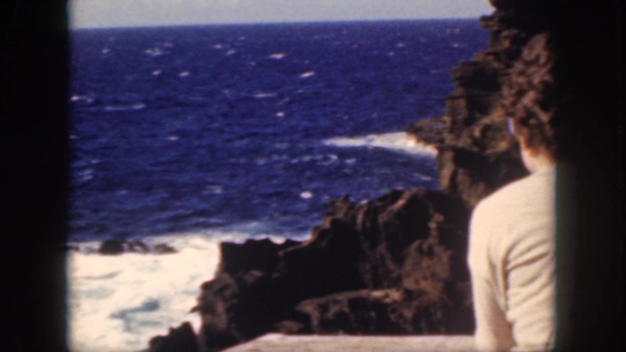 Woman in a pale sweater sits by dark rocks overlooking blue ocean and breaking surf.