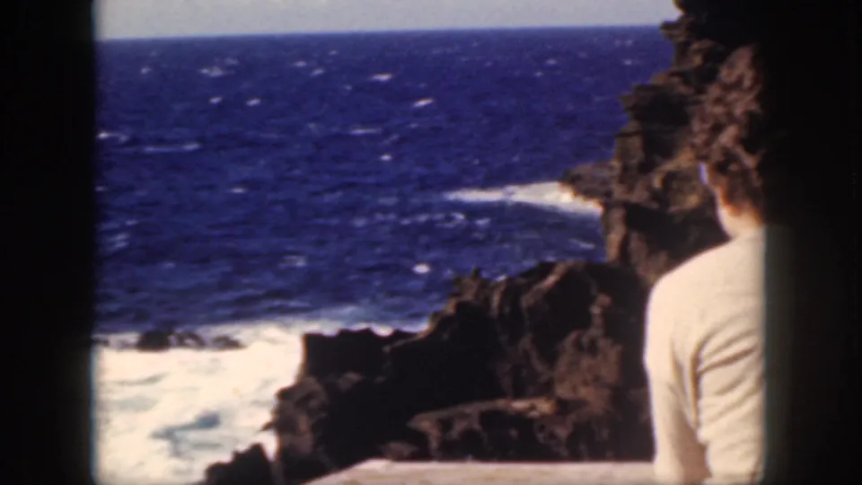 1959 Oahu vacation footage frame with a woman seated beside volcanic rocks, facing the blue ocean and white shoreline surf.