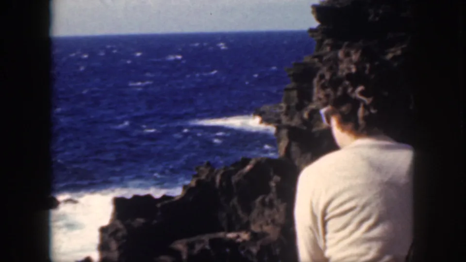 1959 Oahu 8mm frame showing a woman in sunglasses seated near dark coastal rocks, looking out over rough blue water.