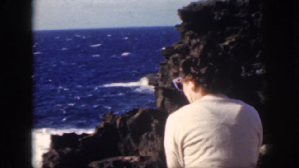 1959 Oahu home-movie still of a woman seated on a cliff edge, framed by dark rocks, blue ocean, and white surf.