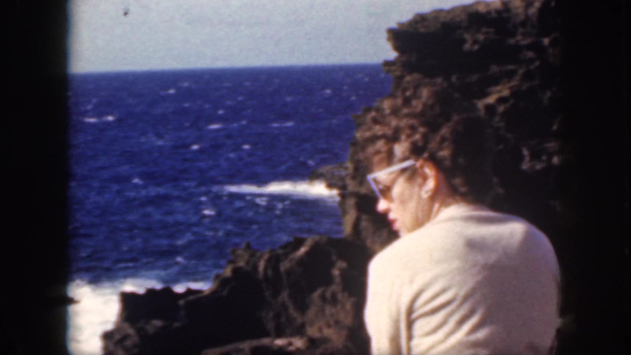 In 1959 Oahu home-movie footage, a woman in white-framed sunglasses sits on volcanic rocks overlooking the blue ocean.