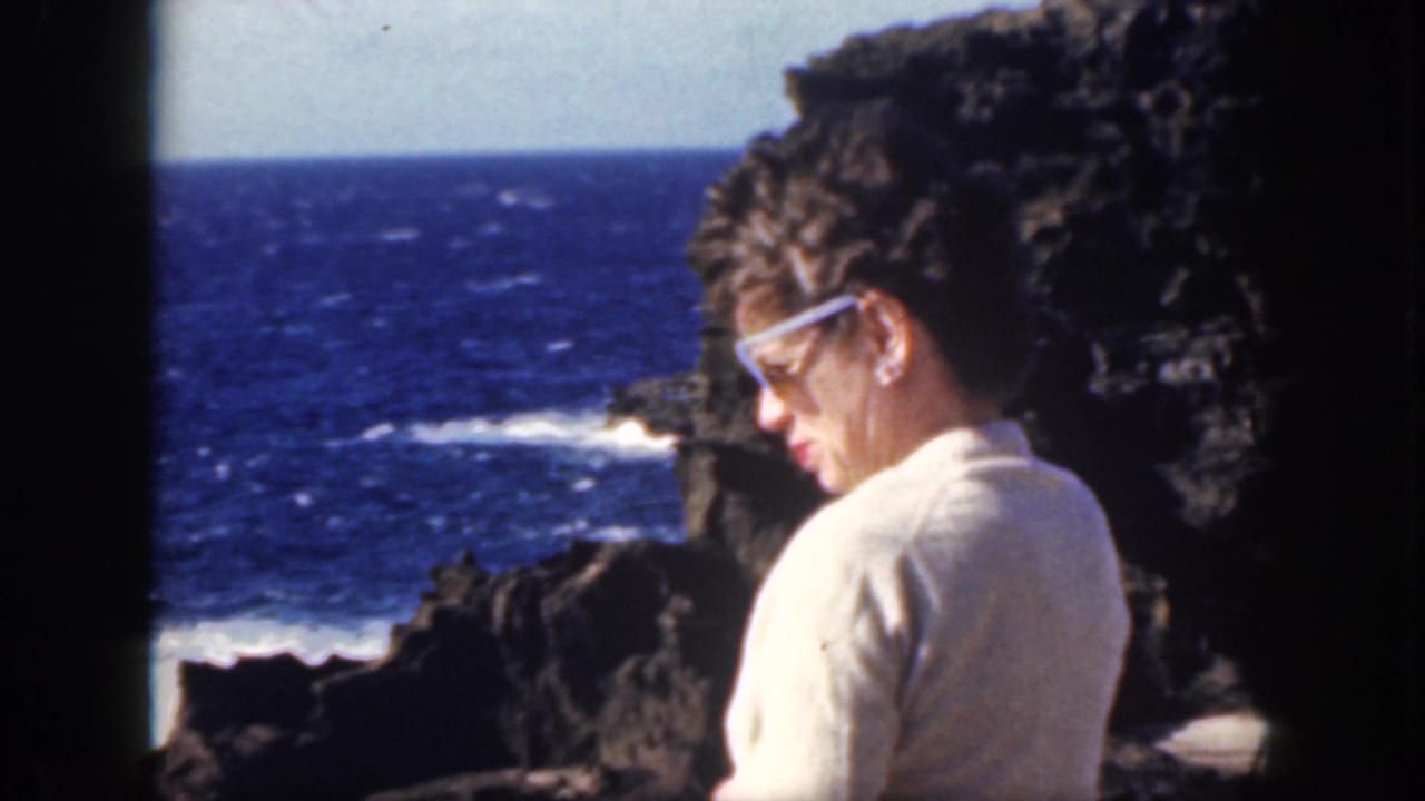 Woman in profile wearing white sunglasses near rocks and breaking surf.