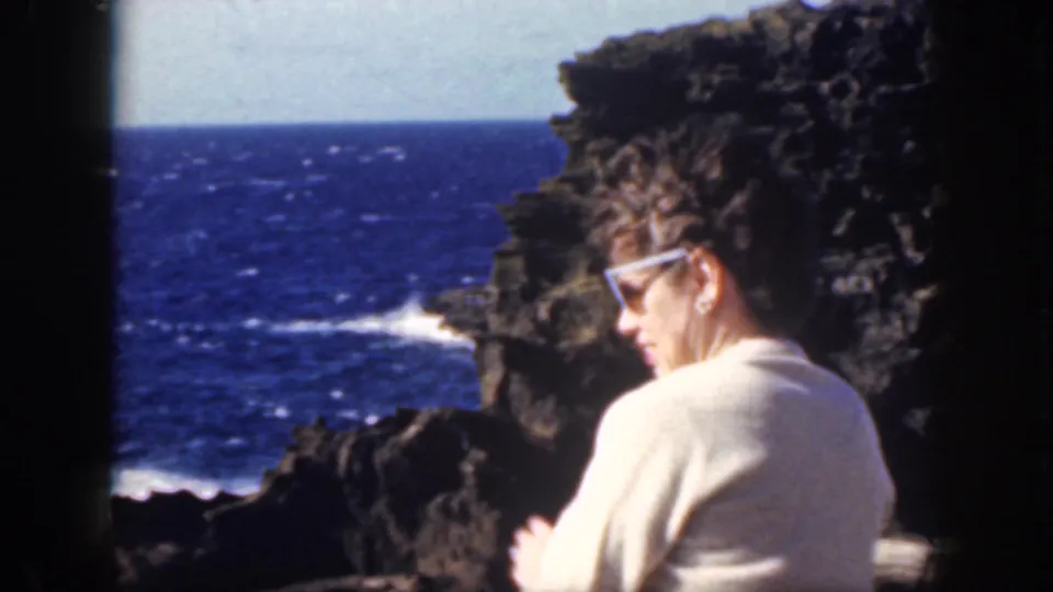 A seated woman in sunglasses looks down near dark lava rocks, with the metadata-supported Oahu ocean shoreline behind her.