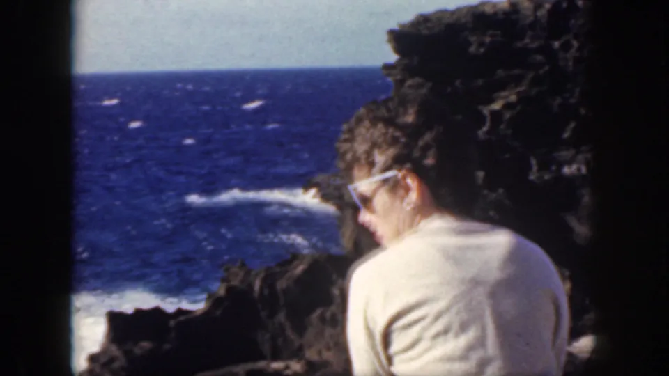 The woman is seen from behind on the rocky Oahu coast, facing deep blue water and white surf along the lava shoreline.