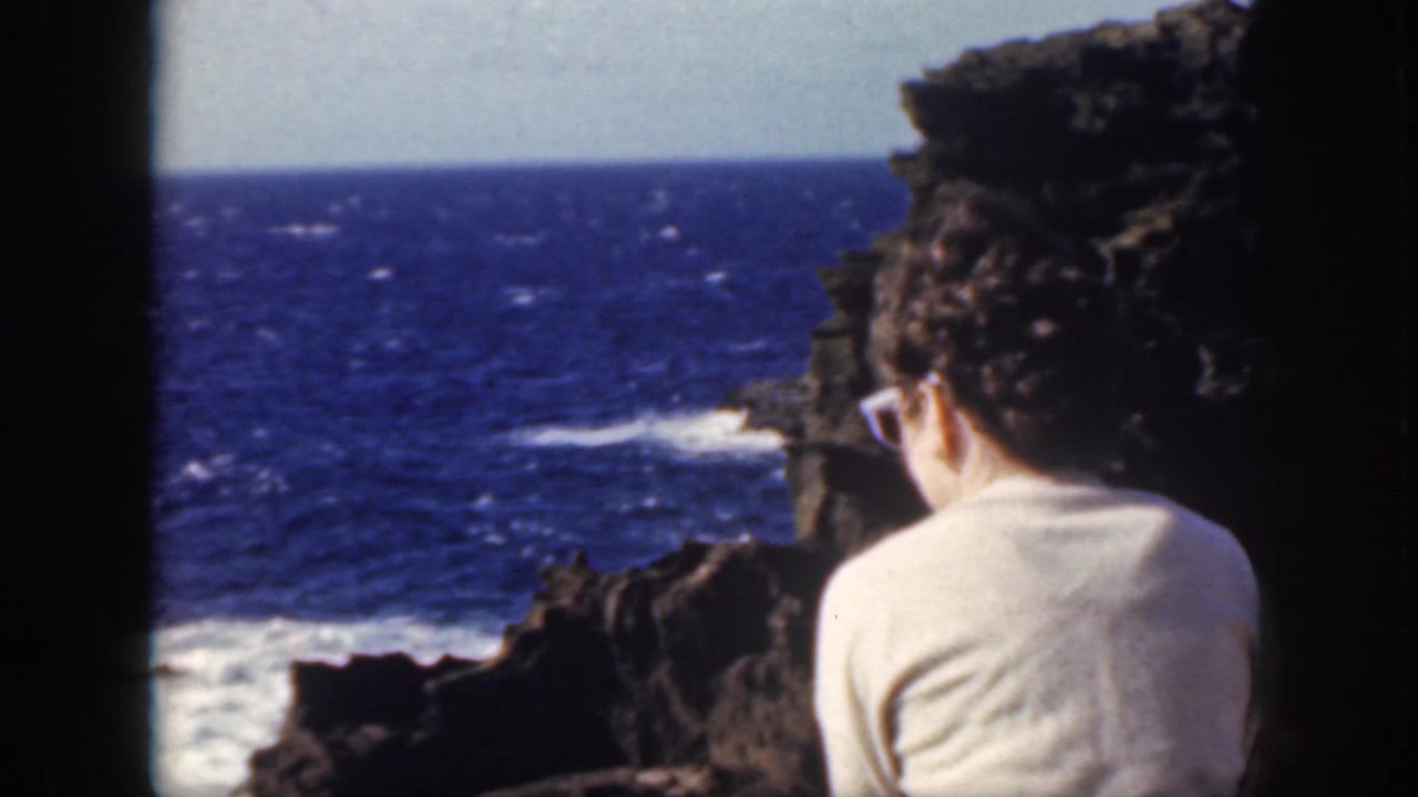 1959 Oahu home-movie frame showing a person from behind beside dark rock, looking toward blue ocean and breaking surf.