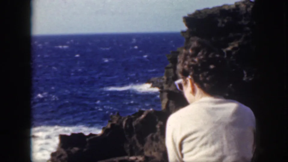 1959 Oahu home-movie frame showing a person from behind beside dark rock, looking toward blue ocean and breaking surf.